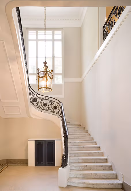 Elegant white marble staircase with ornate black wrought iron railing and hanging chandelier light.