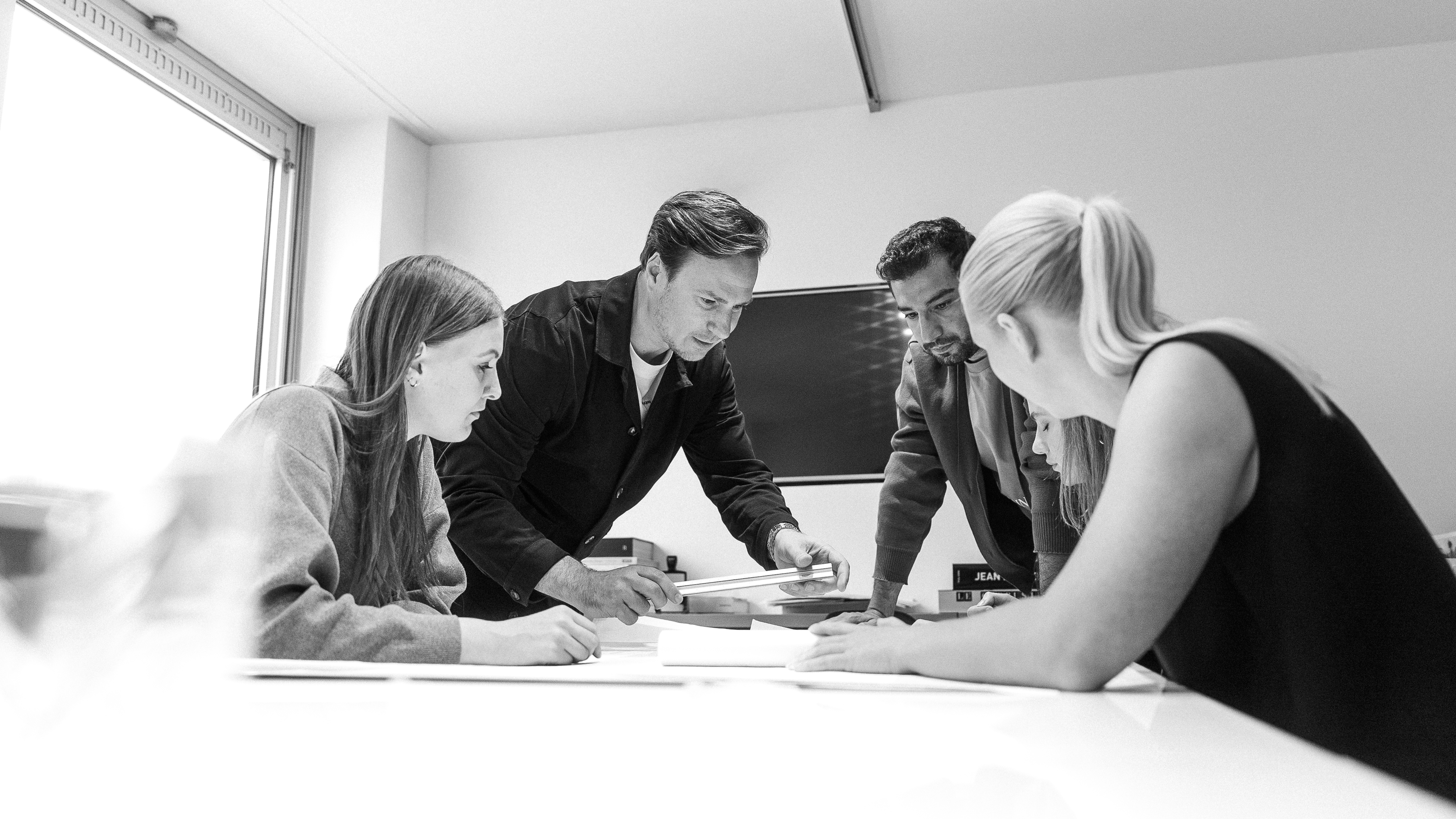 Vier Personen besprechen konzentriert Dokumente an einem Tisch in einem Büro mit Fenster und Monitor.
