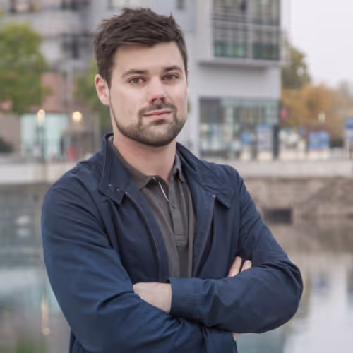 Homme aux cheveux courts et barbe, bras croisés, devant un bâtiment moderne et un plan d’eau.