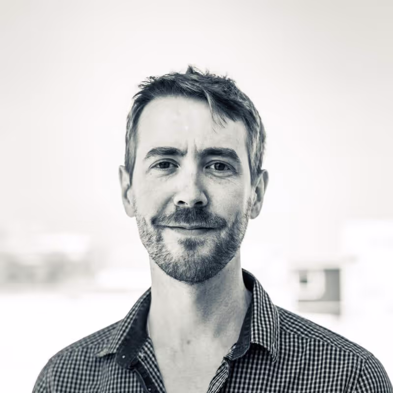 Portrait d'un homme souriant avec barbe légère et chemise à carreaux.