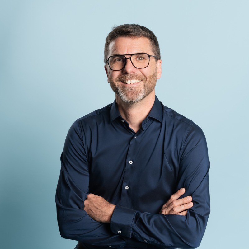 Homme souriant avec des lunettes et chemise bleu marine, bras croisés, fond bleu clair.