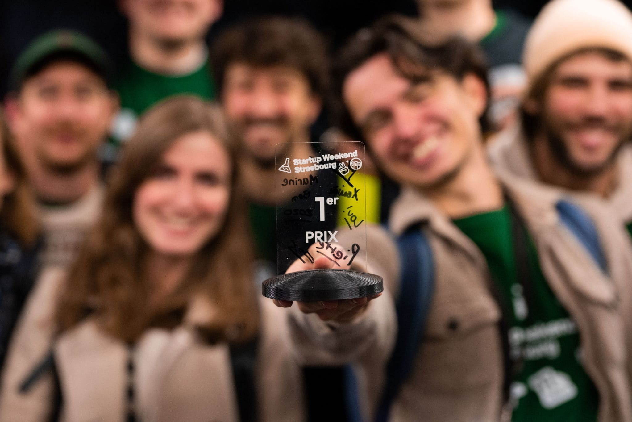 Groupe souriant tenant un trophée de 1er prix Startup Weekend Strasbourg, flou en arrière-plan.