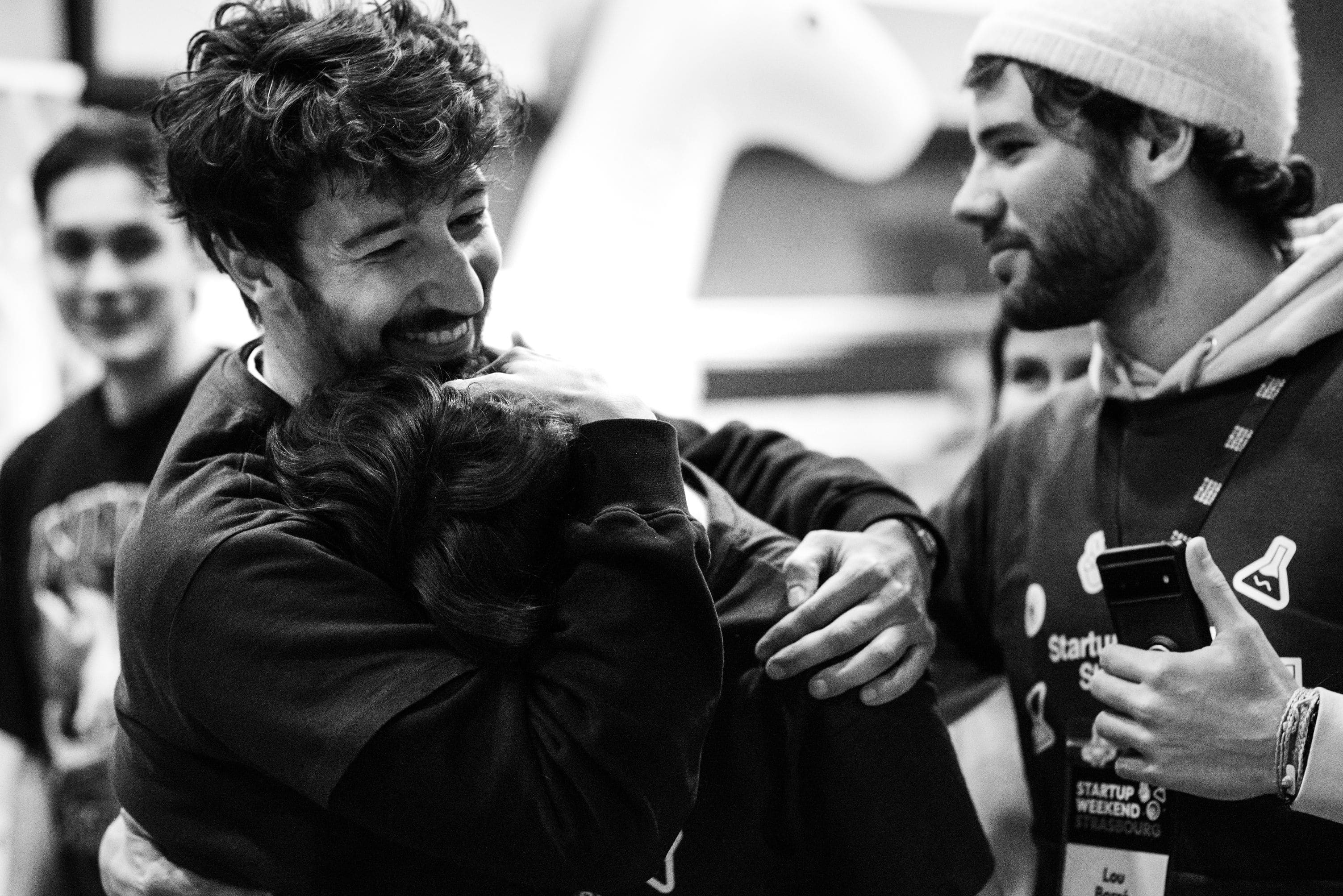 Deux hommes s'enlacent chaleureusement tandis qu'un autre regarde avec un sourire en arrière-plan.