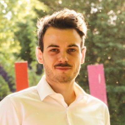 Portrait d'un homme aux cheveux bruns, portant une chemise blanche, avec un fond flou vert et coloré.