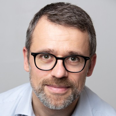 Homme avec lunettes rondes vertes, moustache, chemise blanche, souriant devant un fond noir.