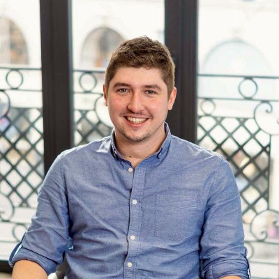 Jeune homme souriant portant un polo bleu sur un fond flou moderne.