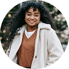 Femme souriante portant un manteau beige et un pull marron clair en extérieur.