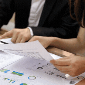 Two people in business attire reviewing and pointing at printed charts and graphs on a table.