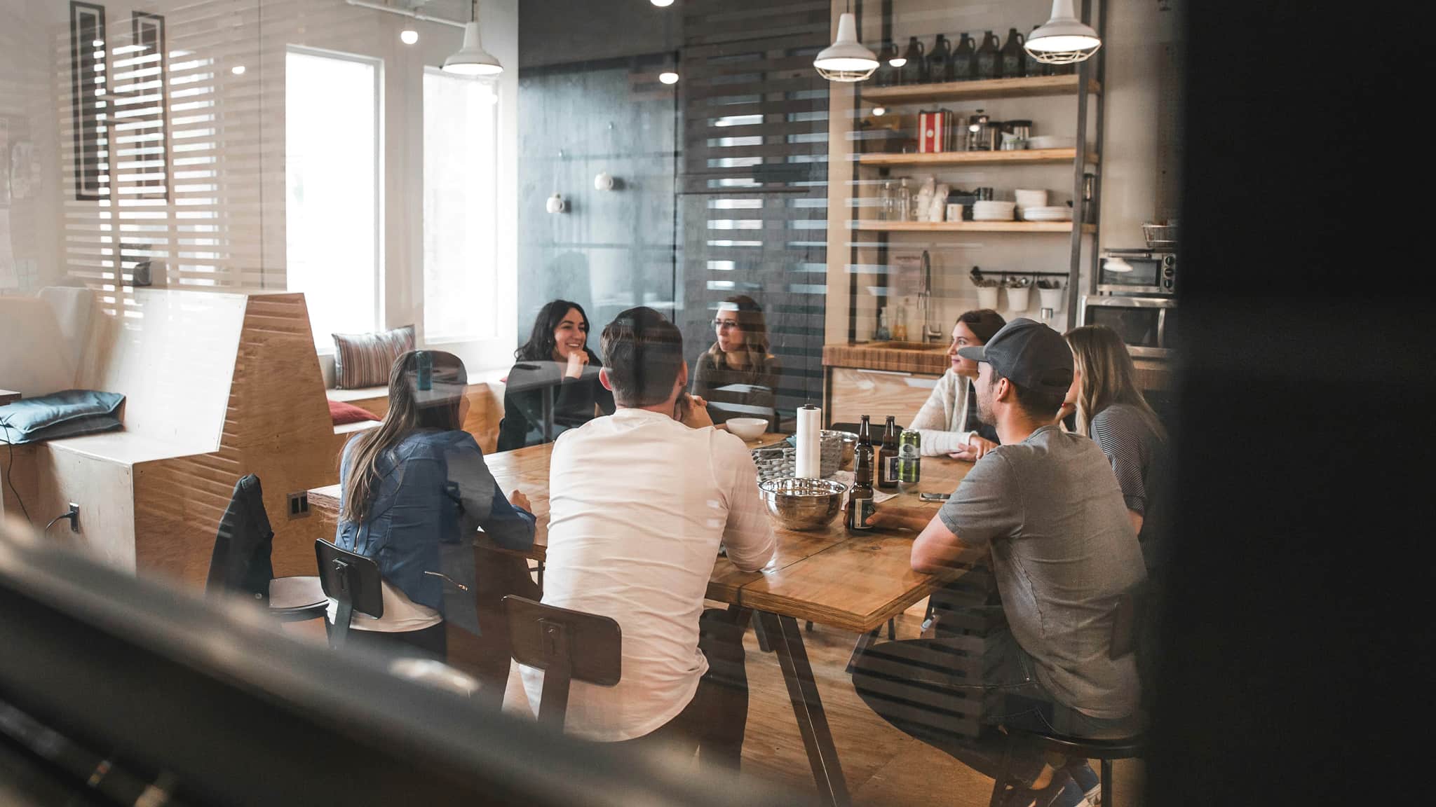 Seven people sitting around a wooden table in a modern kitchen or meeting room, talking and drinking.