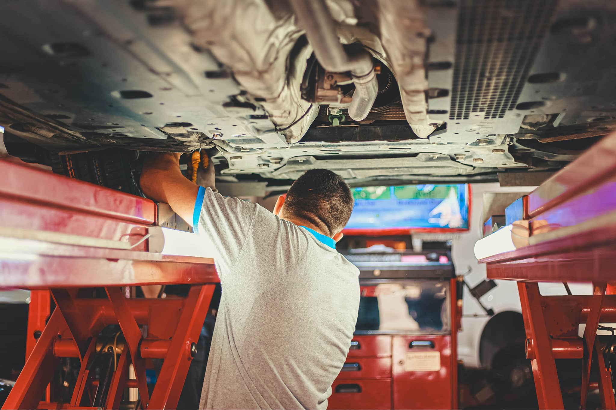 Automotive mechanic in gray shirt working underneath a car lifted on red hydraulic lifts.