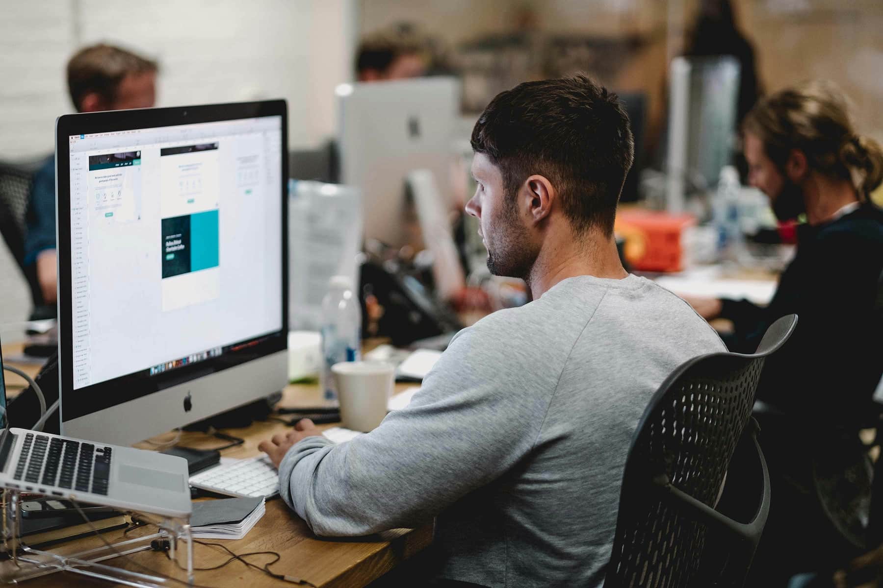 Man in gray shirt working on a desktop computer in a busy office environment with others.