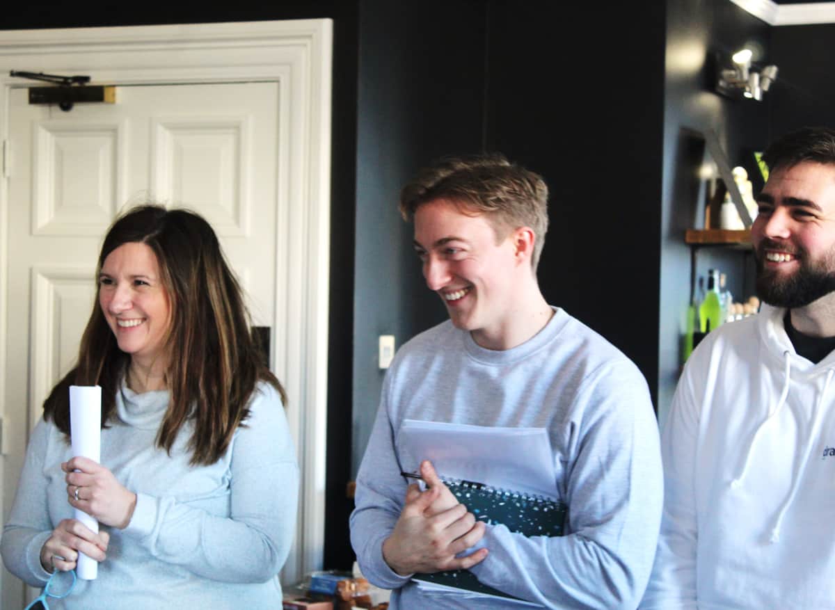 Three people smiling and standing indoors, two holding notebooks and papers, casual clothing.