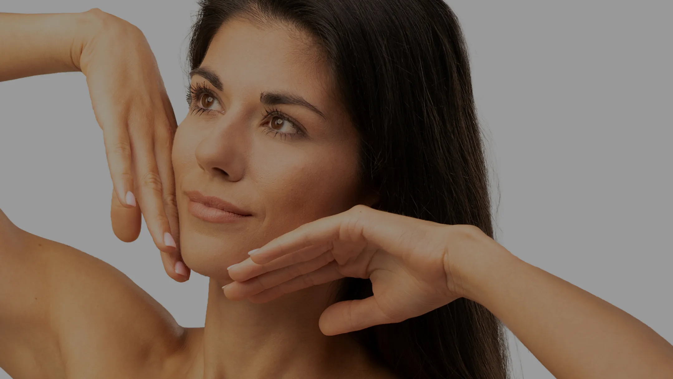 Woman with long dark hair posing with hands framing her face against a simple gray background.