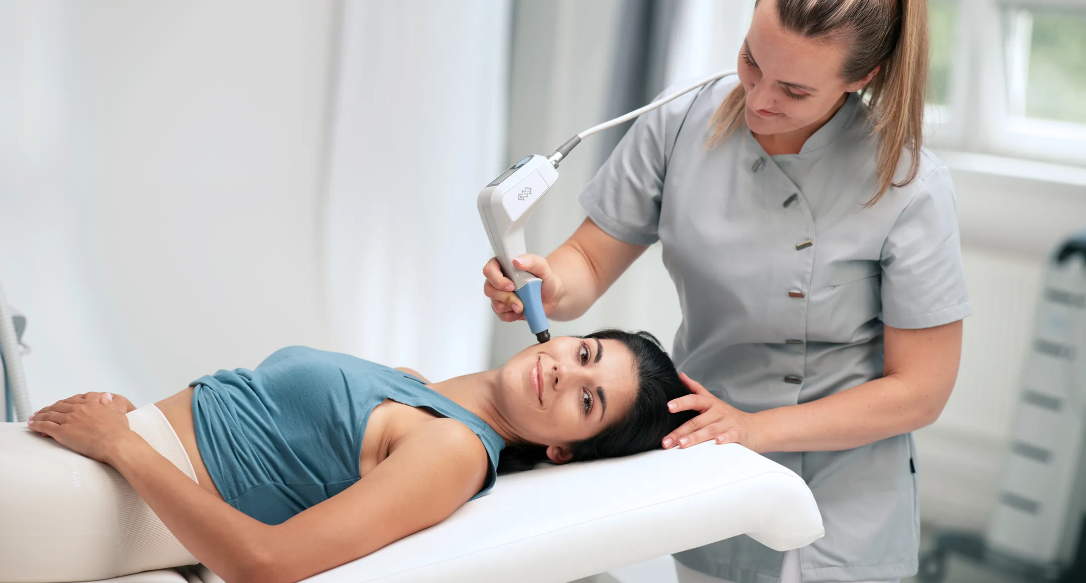 Woman receiving facial treatment with a handheld device from a smiling technician in a clinic.