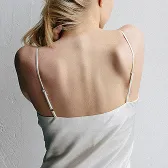 Woman with blonde hair showing bare back wearing a white spaghetti strap top against a white wall.