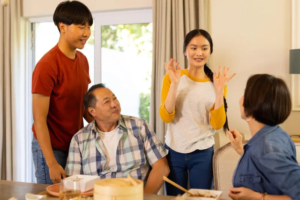 Asian family of four chatting warmly at home around a meal, with a teen girl speaking expressively.