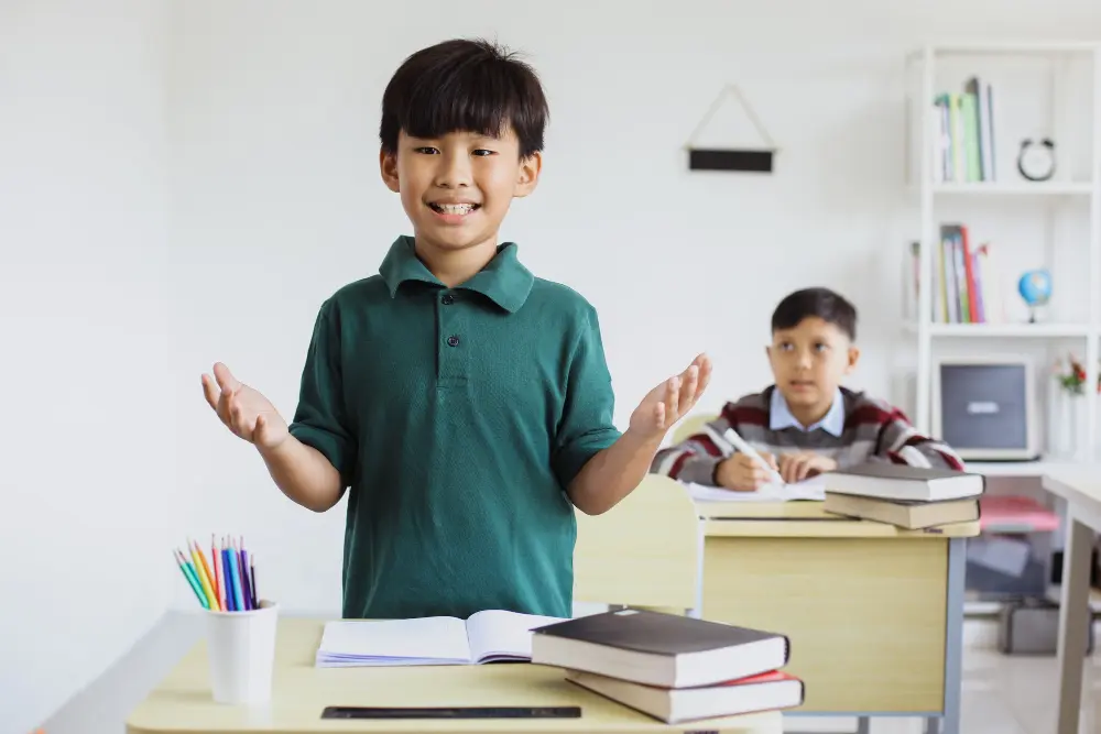 Smiling Asian boy in a green polo shirt presenting with open hands in a classroom, another student at a desk behind him.