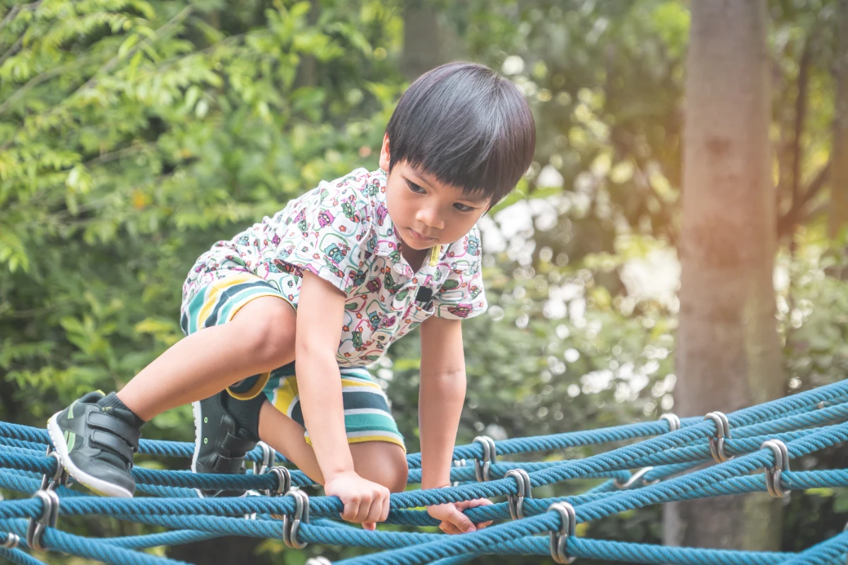 An Asian child in Singapore carefully crossing a playground rope bridge, building confidence by overcoming a challenge