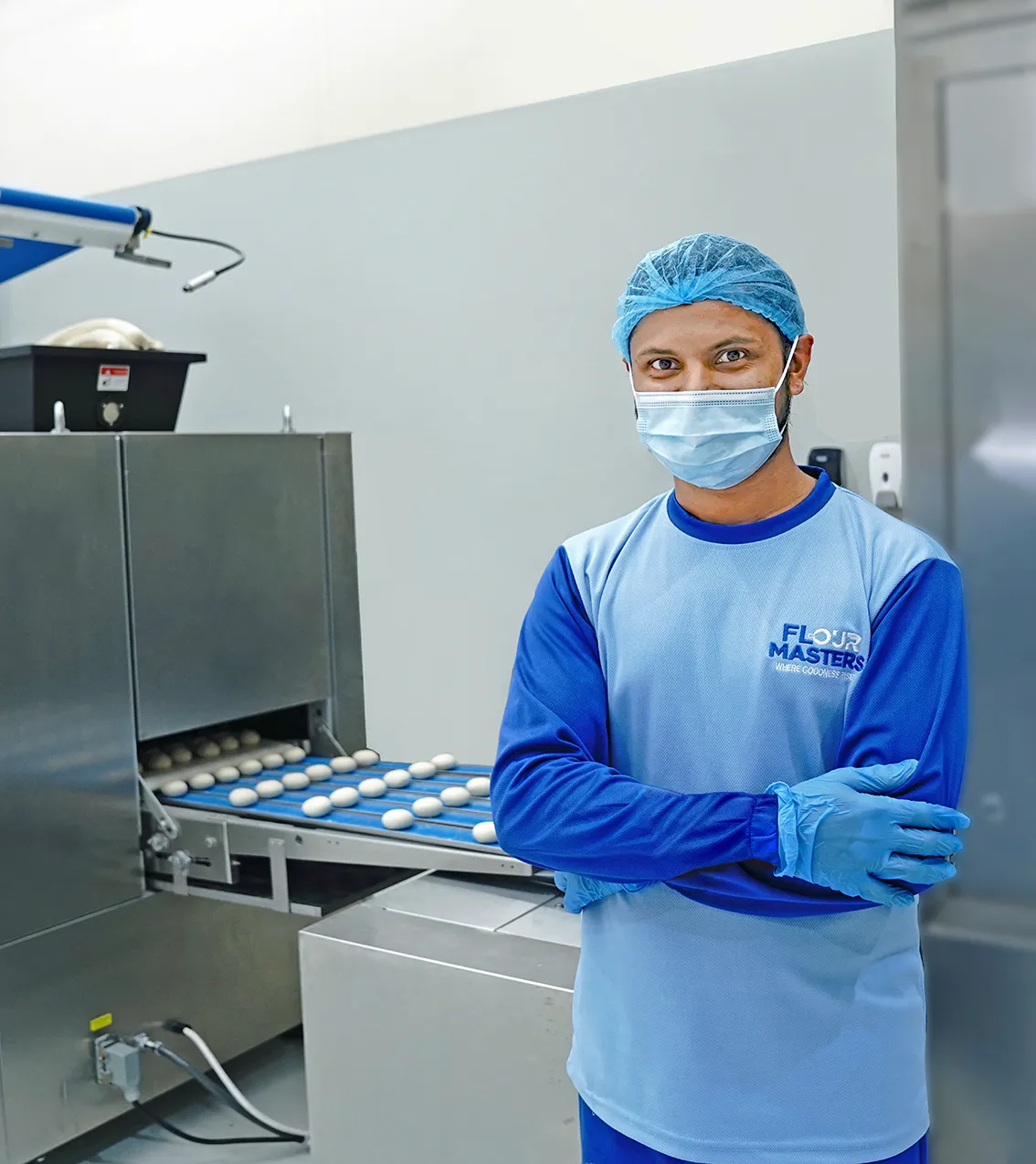 Masked worker in blue uniform and hairnet stands with arms crossed by conveyor with dough balls.