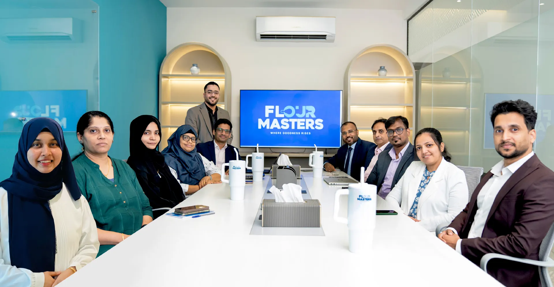 A diverse team of men and women seated around a white conference table with mugs and laptops.