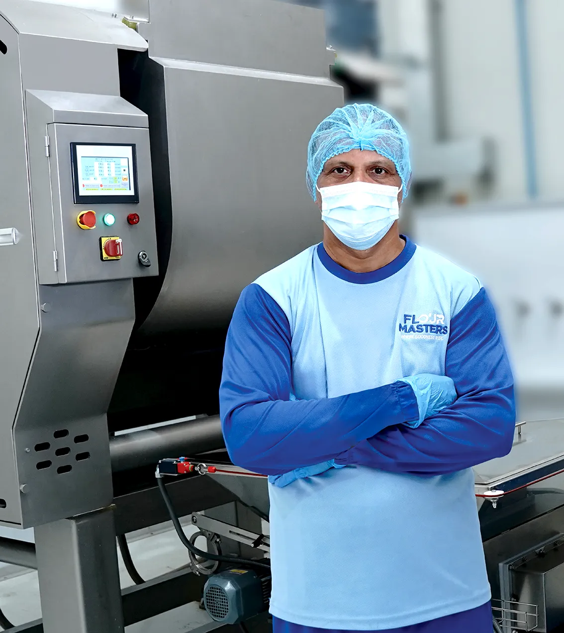 Factory worker in hairnet, mask, gloves, light blue shirt standing with arms crossed by food processing machine.