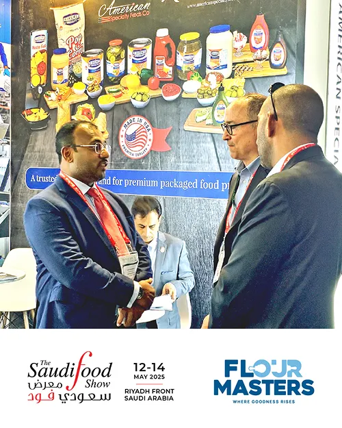 Three men in suits talking at a food trade show booth with packaged food products displayed behind.