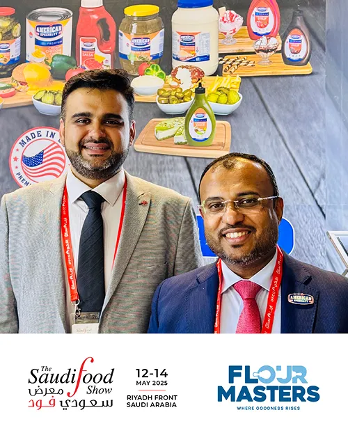 Two men in business attire with red lanyards at Saudi Food Show 2025 with branded food products in background.