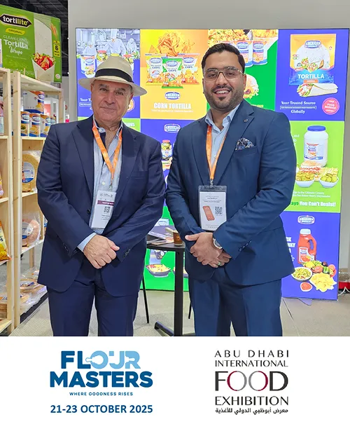 Two men in suits with conference badges stand in front of a colorful food exhibit at Abu Dhabi Food Exhibition.