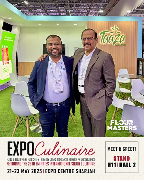 Two men in business attire smiling and posing at the Taaza booth in an expo with white chairs and green carpet.