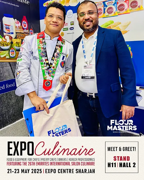 Two men smile at a food expo, one in a chef's uniform holding a Flour Masters tote bag.