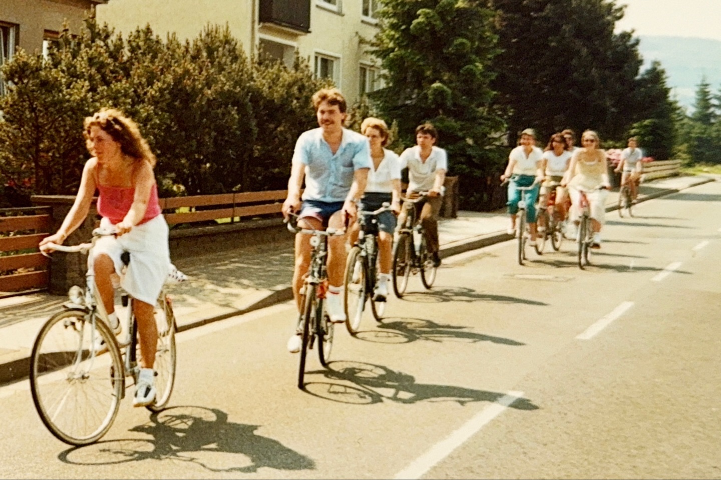 Eine Gruppe von neun Personen fährt auf einer Straße in einer ländlichen Umgebung Fahrrad, begleitet von Bäumen und Häusern im Hintergrund, an einem sonnigen Tag.