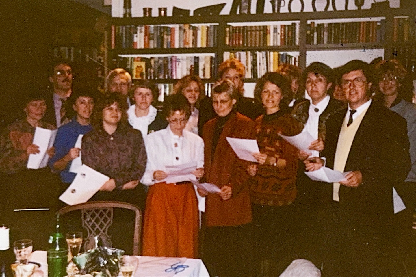 Gruppe von 15 Personen, die drinnen vor einem Bücherregal stehen und Papierblätter in den Händen halten, vermutlich zum gemeinsamen Lesen oder Singen bei einer Veranstaltung.