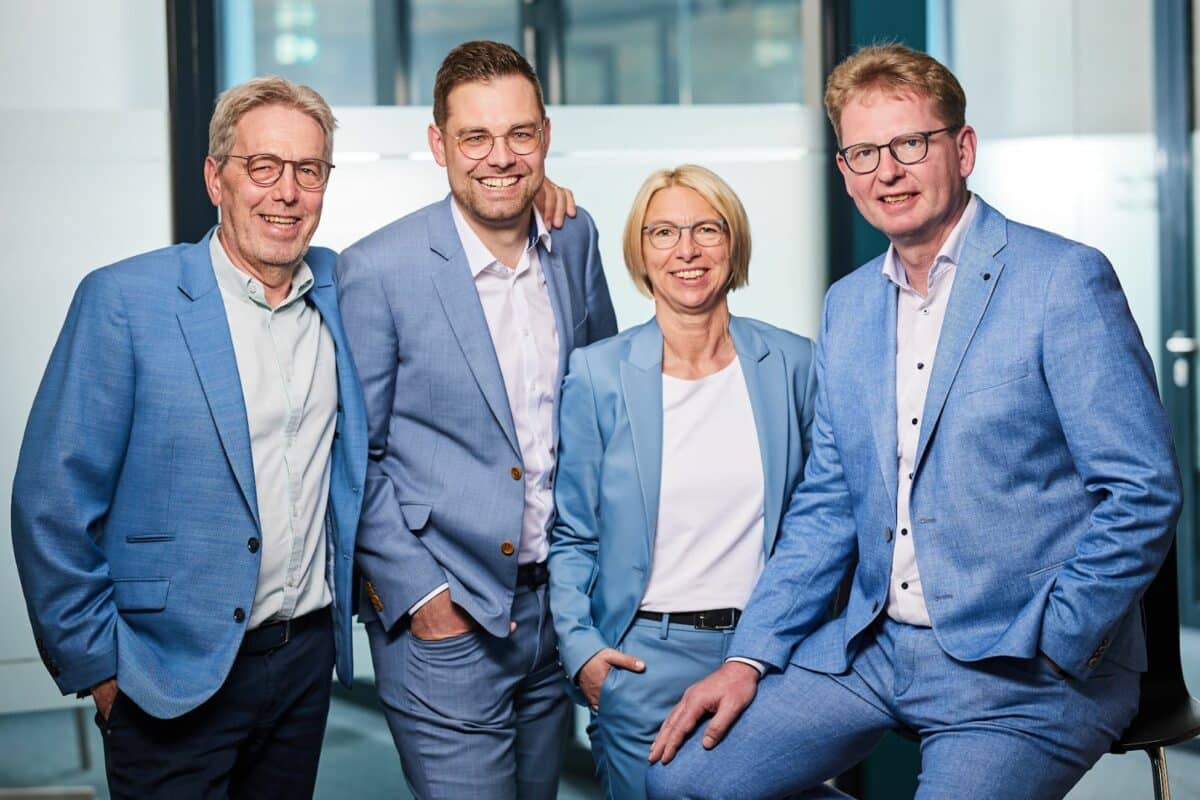 Vier lächelnde Personen, drei Männer und eine Frau, alle tragen hellblaue Anzüge und Brillen, posieren zusammen im Büro, zwei Männer stehen, die andere sitzt auf einem Hocker.