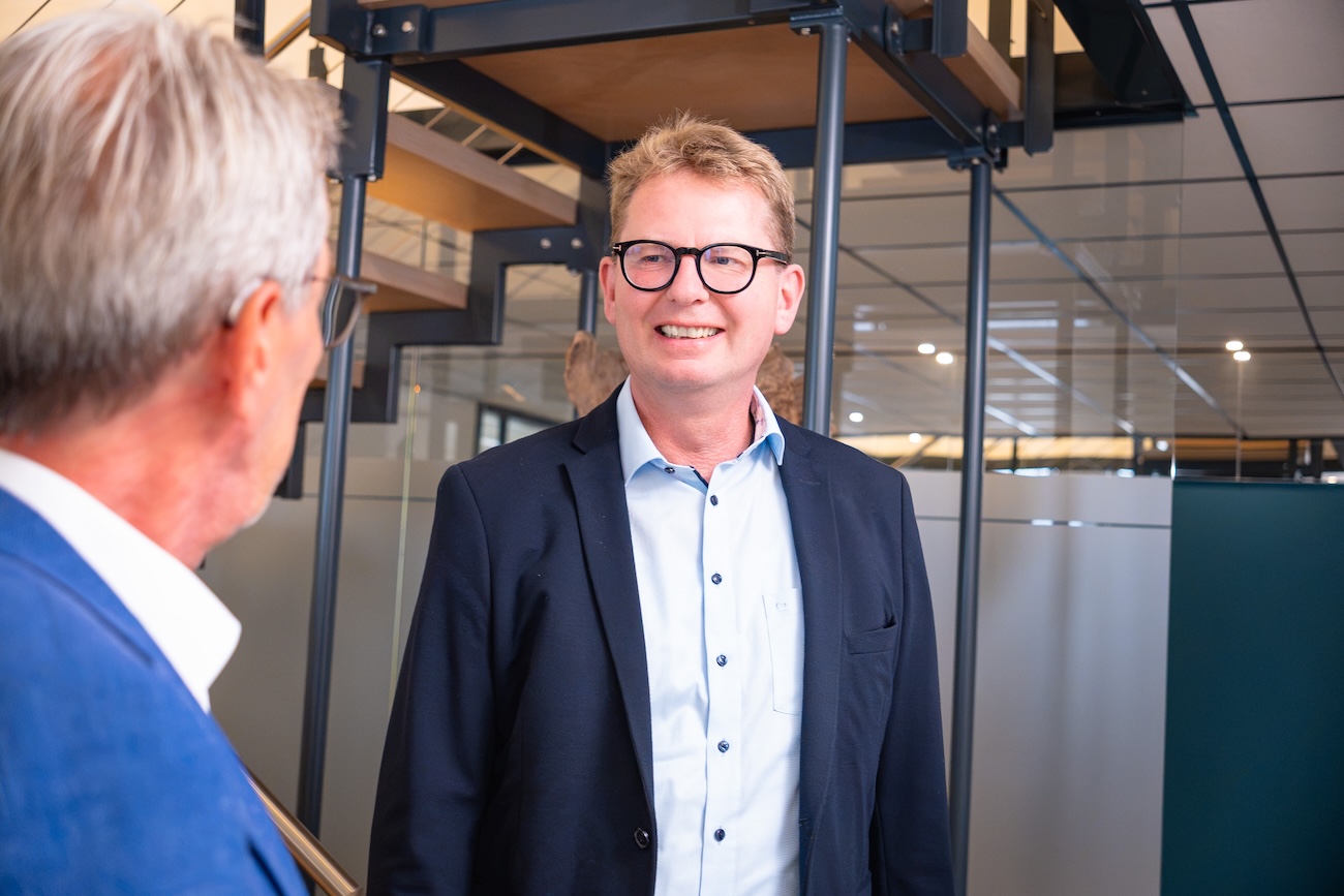 Zwei Männer in formeller Kleidung sprechen in einem modernen Büro mit Glaswänden und einer Treppe im Hintergrund. Der Mann im Vordergrund trägt Brille, blaue Hemd und schwarzen Blazer und lächelt.