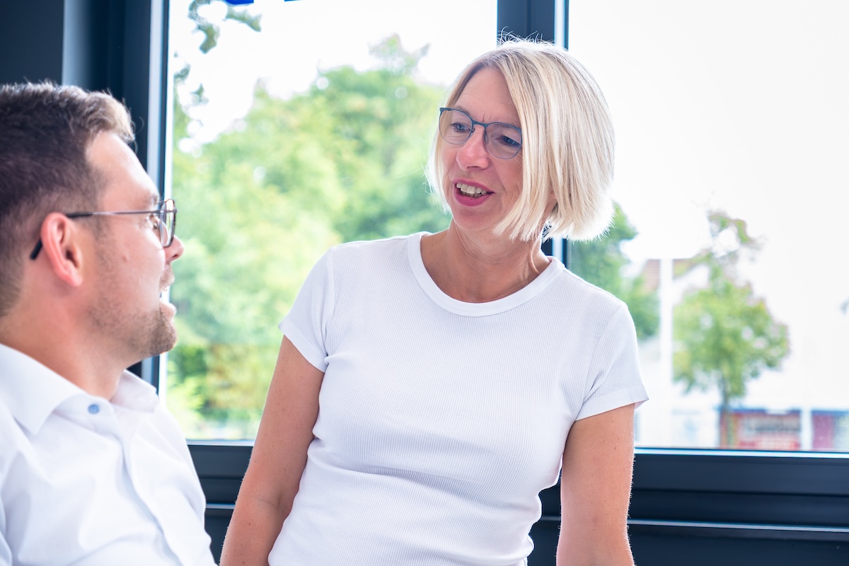 Eine lächelnde Frau mit blonden, kinnlangen Haaren und einer weißen Bluse unterhält sich mit einem Mann mit kurzen braunen Haaren und Brille vor einem Fenster mit Blick auf grüne Bäume.