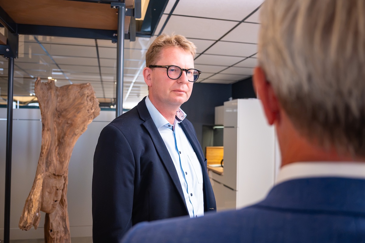 Ein Mann mit Brille, hellblauem Hemd und dunklem Sakko steht in einem modernen Büro vor einem anderen Mann mit grauem Haar und blauem Sakko, der im Vordergrund unscharf ist.