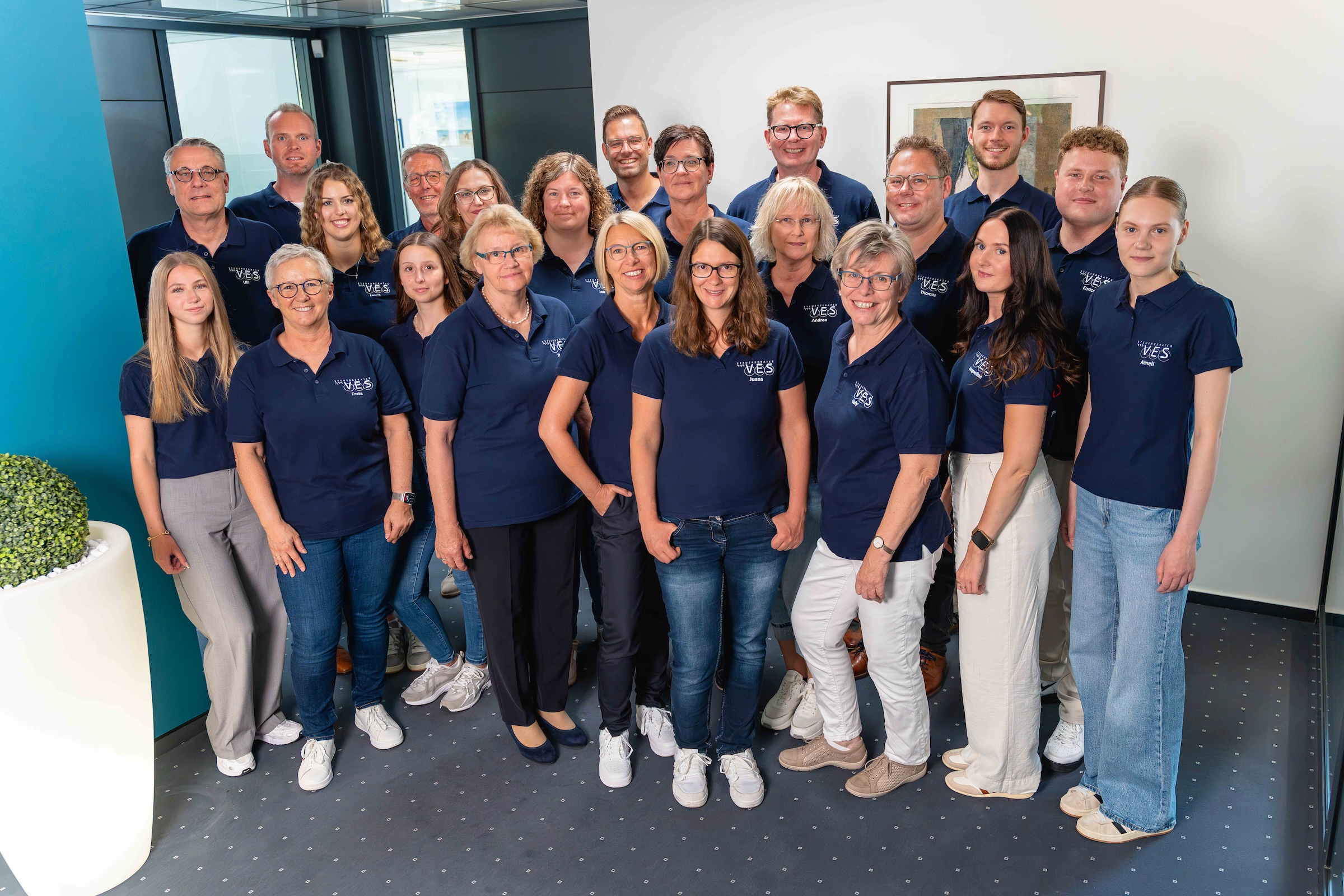 Gruppenfoto von 22 Personen, die in einem Innenraum stehen, alle tragen dunkelblaue Poloshirts mit einem weißen Logo auf der linken Brust, lächeln in die Kamera, hinter ihnen ist eine weiße Wand mit einem gerahmten Bild und links ein modernes blaues Bürointerieur mit einer großen weißen Pflanzenschale.