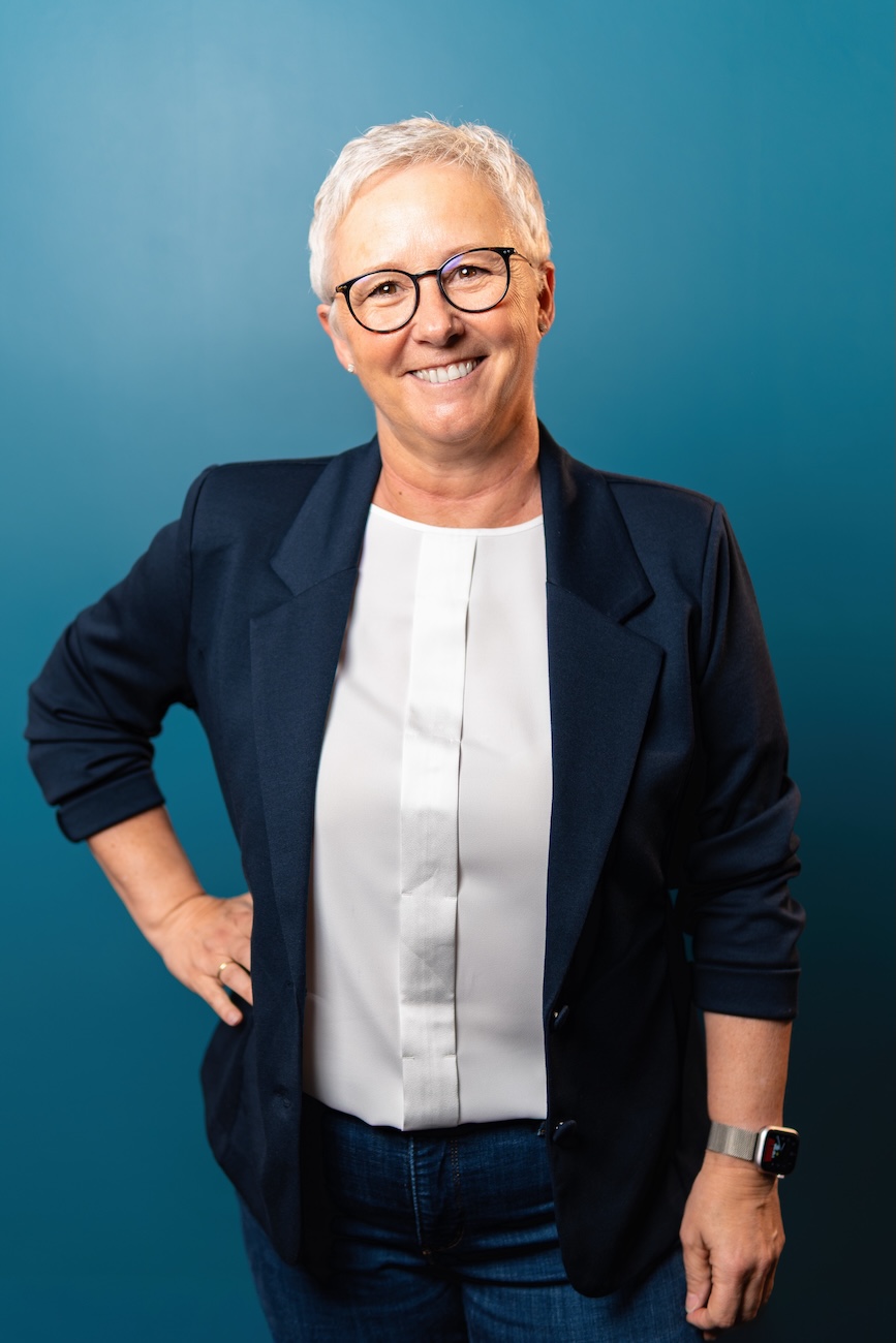 Frau mit kurzen weißen Haaren und runder Brille lächelt freundlich, trägt dunkelblaue Jacke über weißer Bluse und Jeans, steht vor blauem Hintergrund.