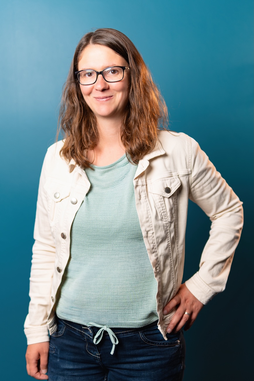 Porträt einer lächelnden Frau mit mittellangen braunen Haaren und Brille, die eine weiße Jeansjacke und ein hellgrünes Oberteil trägt, vor einem blauen Hintergrund stehend, mit einer Hand in der Hosentasche und der anderen am Hüftgürtel.