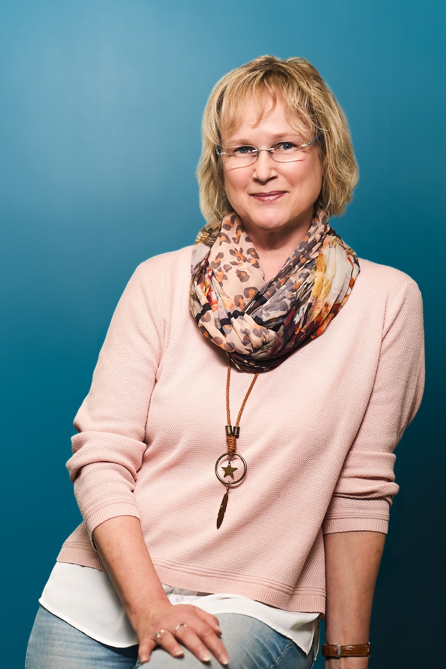 Porträt einer lächelnden Frau mit kurzen blonden Haaren, die eine Brille, einen gemusterten Schal, einen rosa Pullover, eine Halskette mit Stern und Feder sowie eine Uhr trägt vor einem blauen Hintergrund.