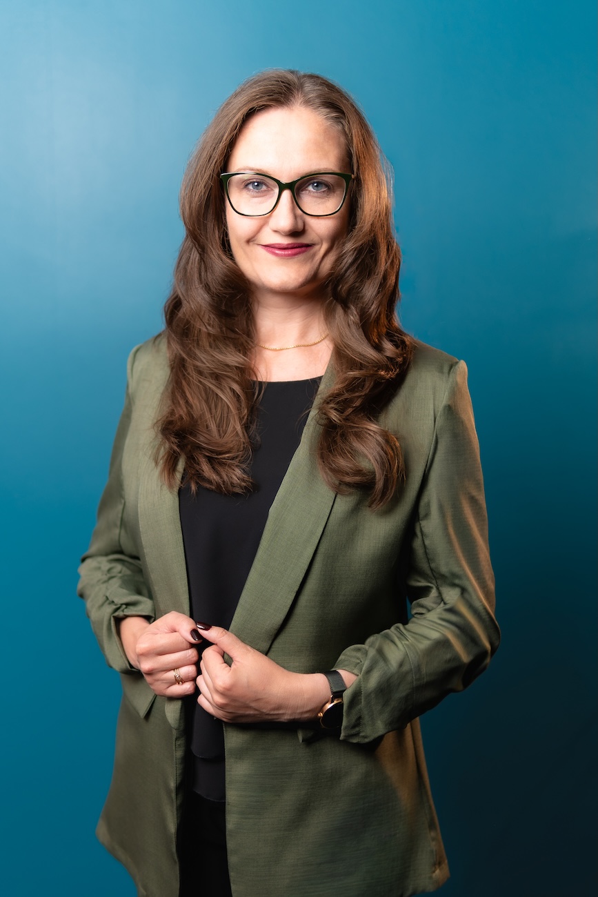 Porträt einer Frau mit langen braunen Haaren und Brille, die eine grüne Jacke und ein schwarzes Oberteil vor einem blauen Hintergrund trägt und leicht lächelt.