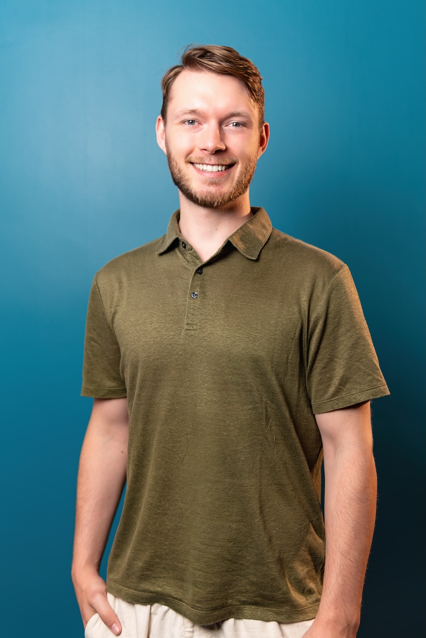 Junger Mann mit kurzem braunem Haar und Bart, der ein olivgrünes Poloshirt und helle Hose trägt, vor einem blauen Hintergrund lächelnd.
