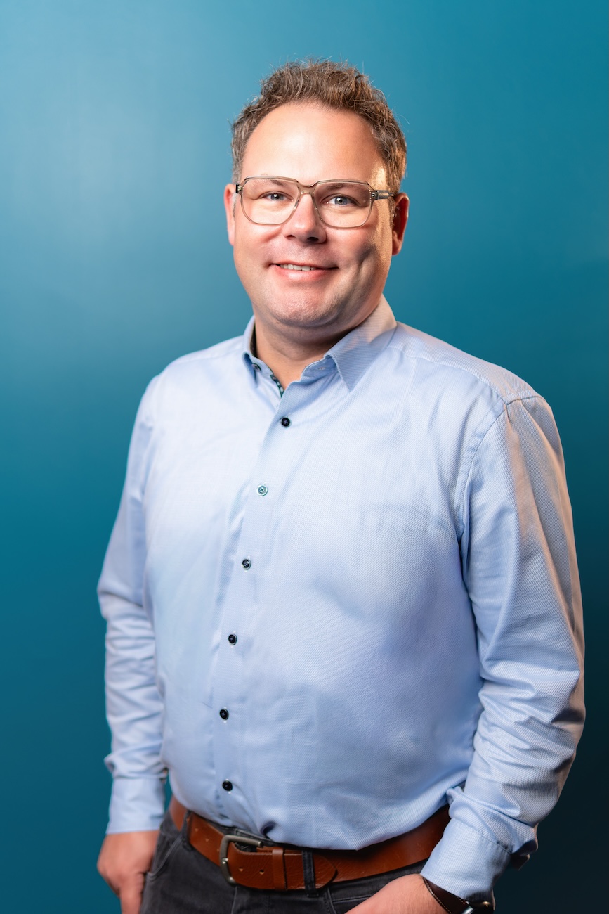 Porträt eines lächelnden Mannes mit kurzen braunen Haaren und Brille, der ein hellblaues Hemd, braunen Gürtel und Armbanduhr trägt vor blauem Hintergrund.