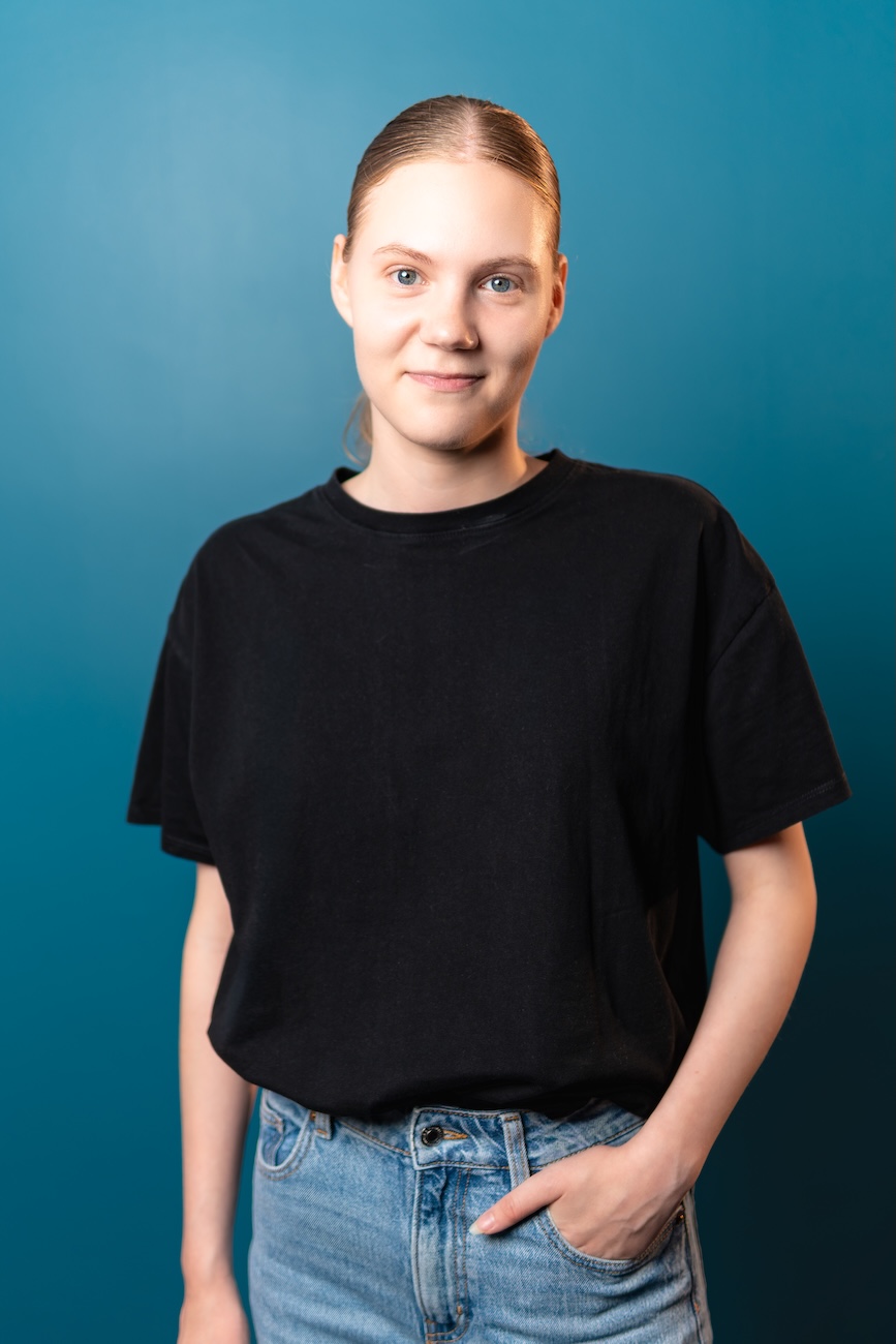 Junge Frau mit blondem, zurückgebundenem Haar trägt ein schwarzes T-Shirt und blaue Jeans vor einem einfarbigen blauen Hintergrund, sie blickt freundlich in die Kamera mit einer Hand in der Hosentasche.