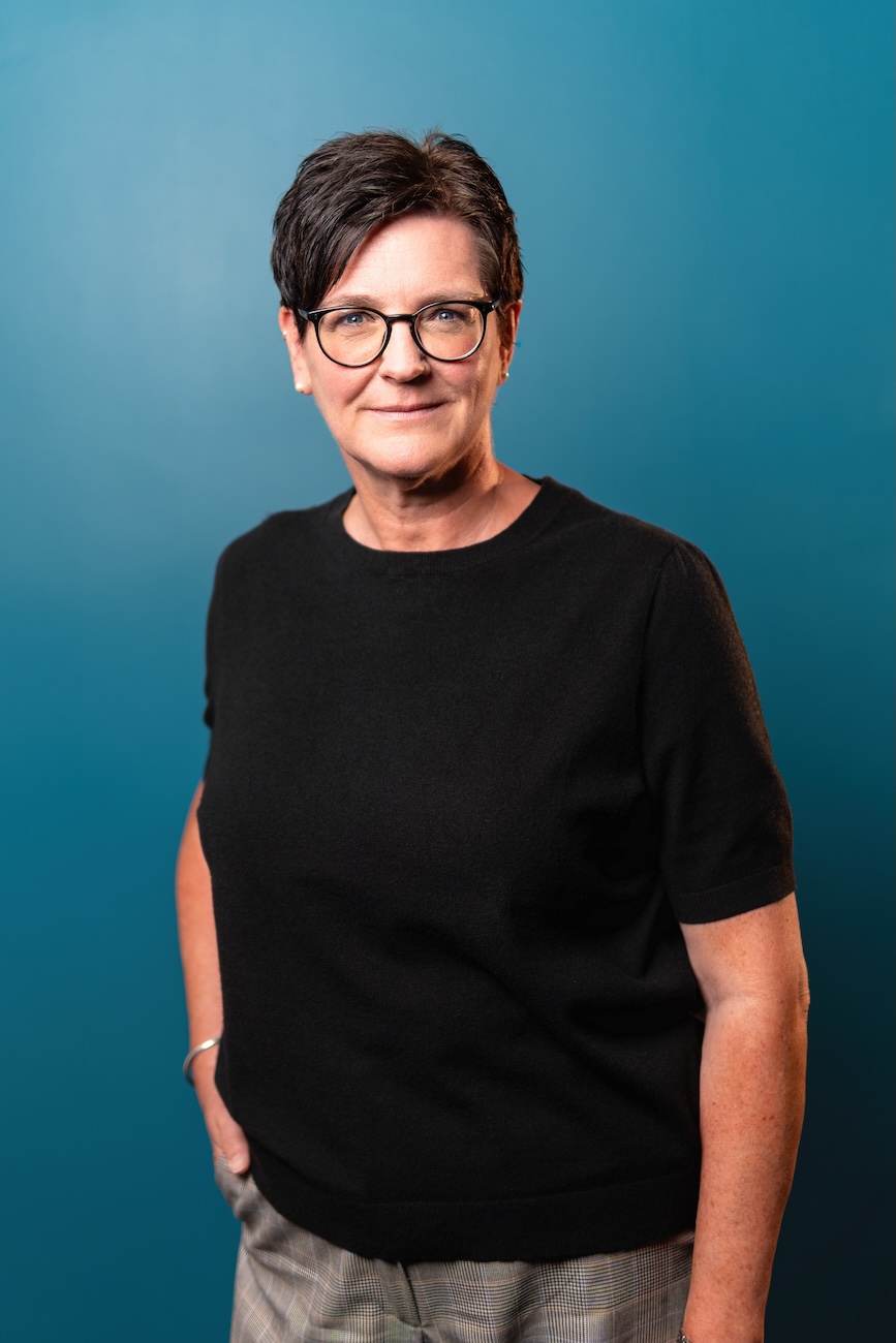 Porträt einer Frau mit kurzen dunklen Haaren und Brille, die ein schwarzes kurzärmeliges Oberteil und karierten Hosen trägt, vor einem blauen Hintergrund.