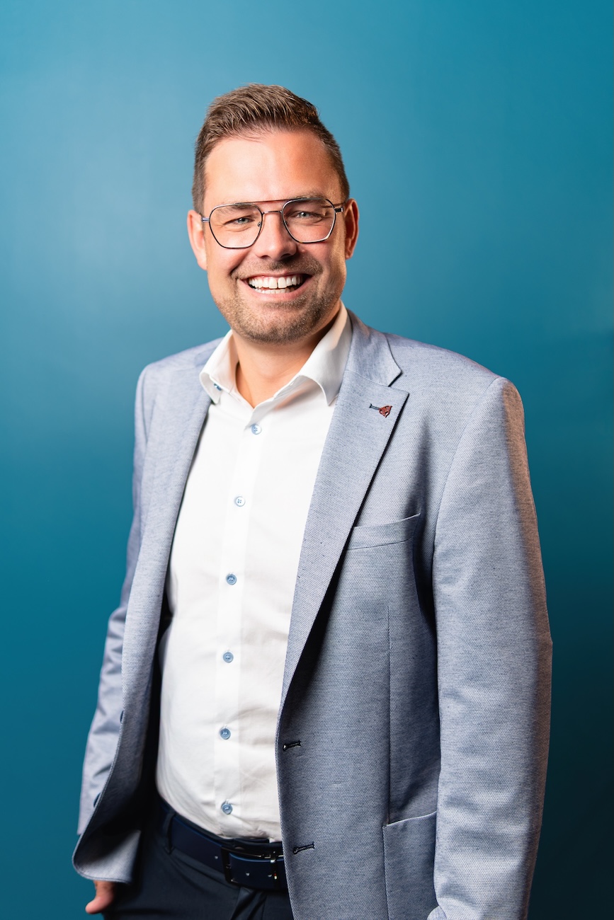 Lächelnder Mann mit Brille trägt helles Sakko, weißes Hemd und dunkle Hose vor blauem Hintergrund.
