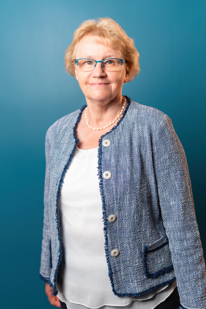 Porträt einer älteren Frau mit blonden, kurzen Haaren, die blaue Brille, eine Perlenkette, eine weiße Bluse und eine blaue Jacke trägt, vor einem einfarbigen blauen Hintergrund.