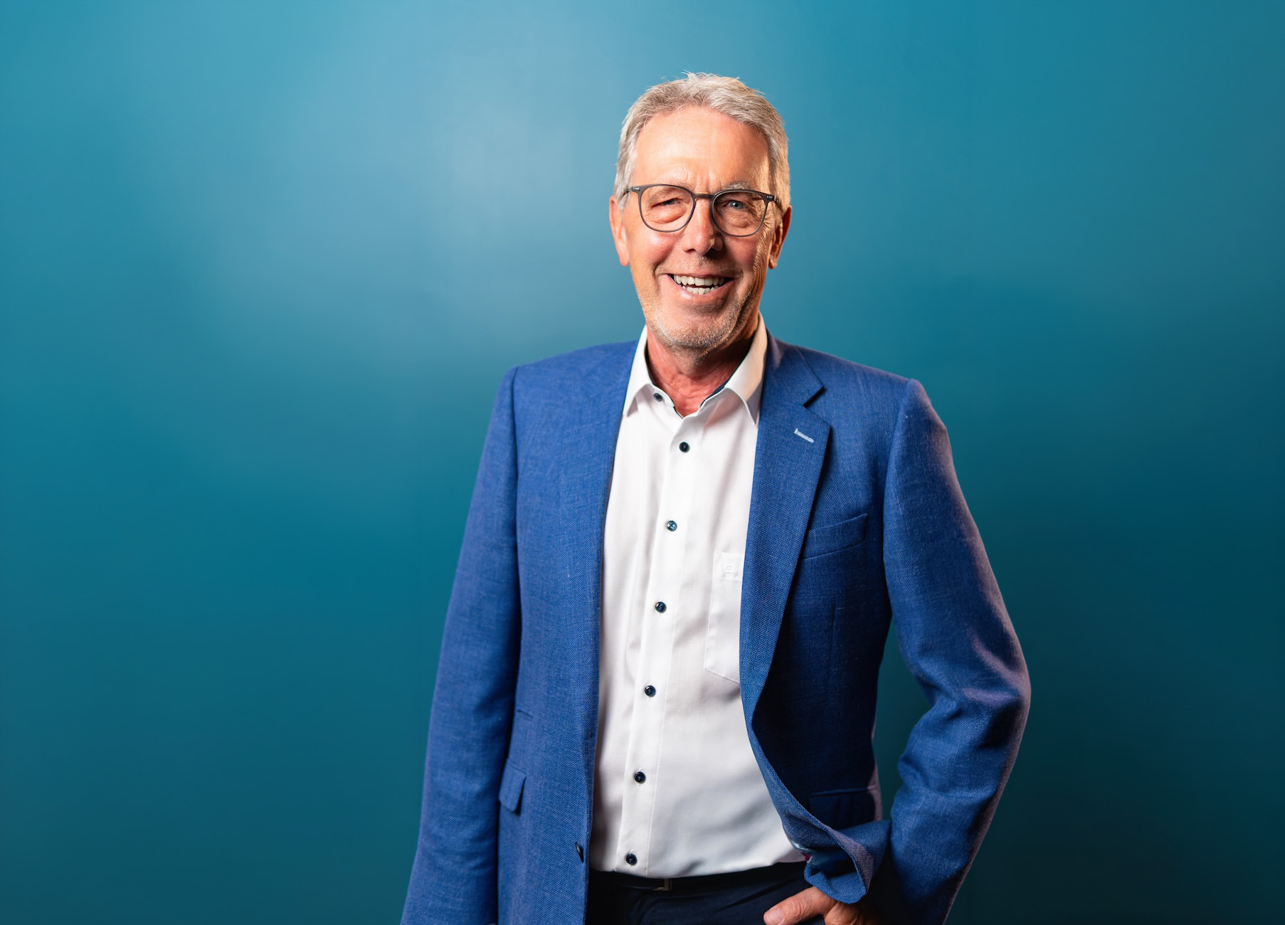 Älterer Mann mit grauem Haar und Brille, der lächelnd in die Kamera schaut, trägt ein blaues Sakko über einem weißen Hemd vor einem einfarbigen blau-grünen Hintergrund.