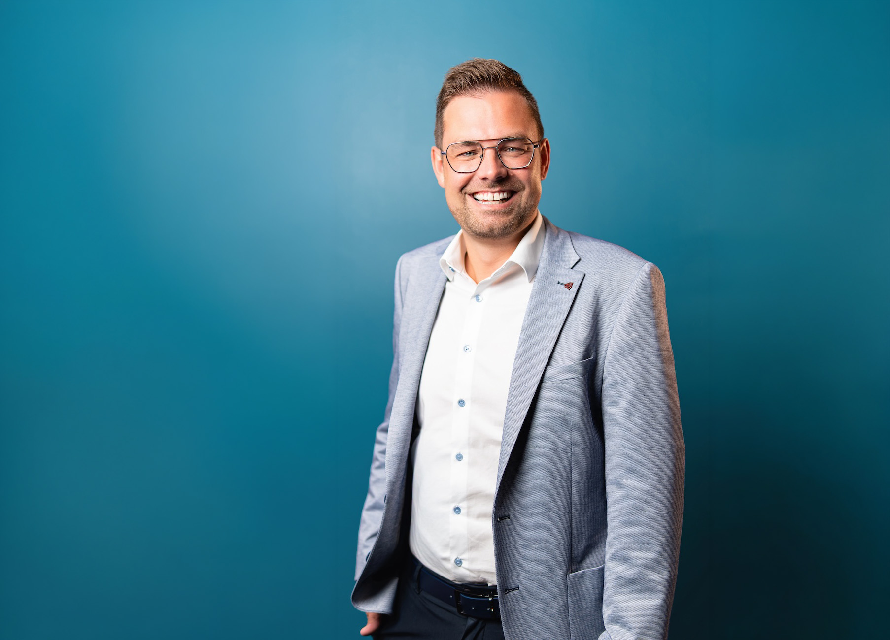 Fröhlicher Mann mit hellgrauem Sakko, weißem Hemd und Brille steht vor blauem Hintergrund.