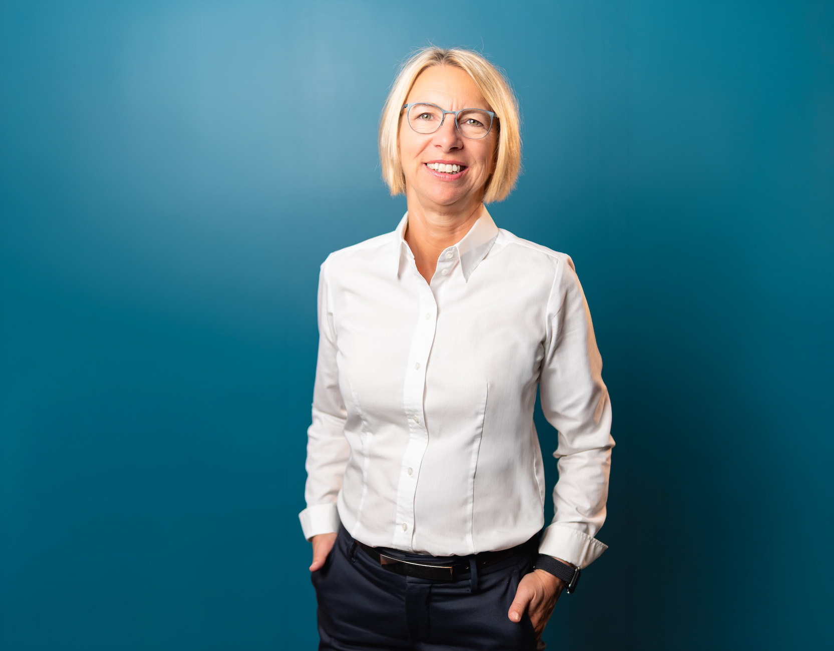 Lächelnde Frau mit kurzen blonden Haaren, die eine weiße Bluse, schwarze Hose und Brille trägt, vor blauem Hintergrund.